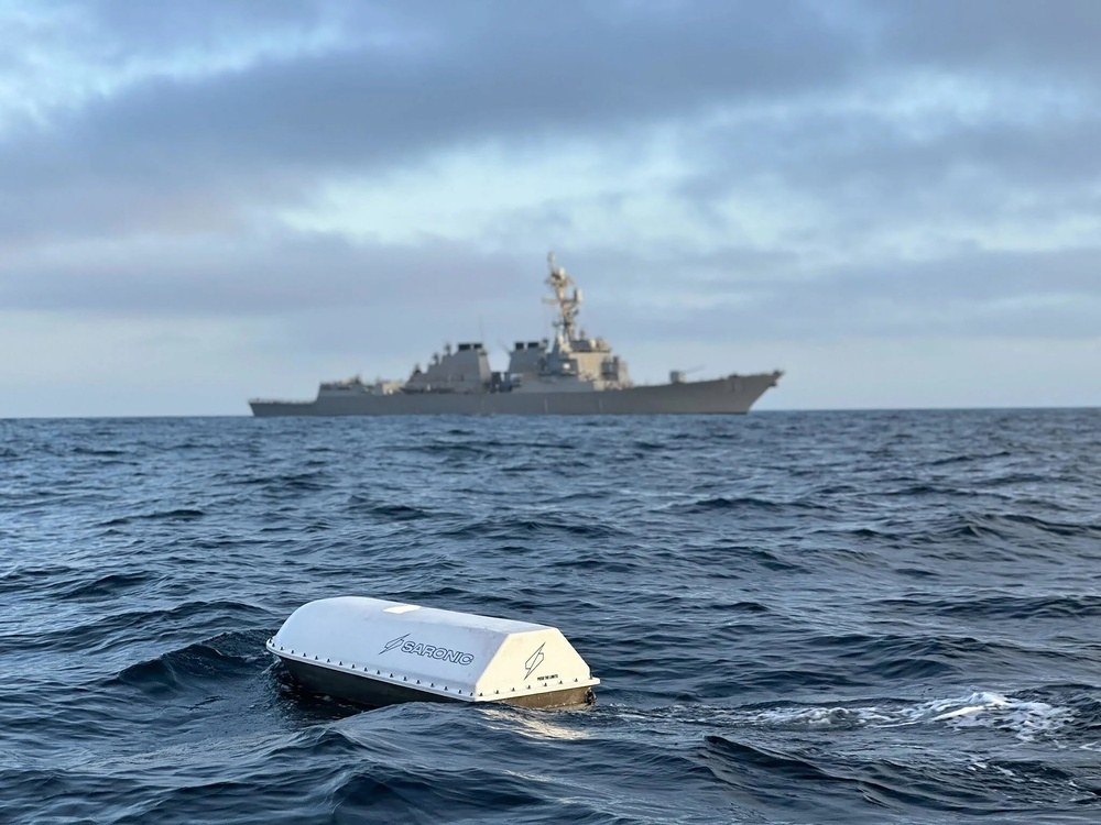 A white Saronic unmanned surface vessel in the open ocean with a Navy destroyer behind it