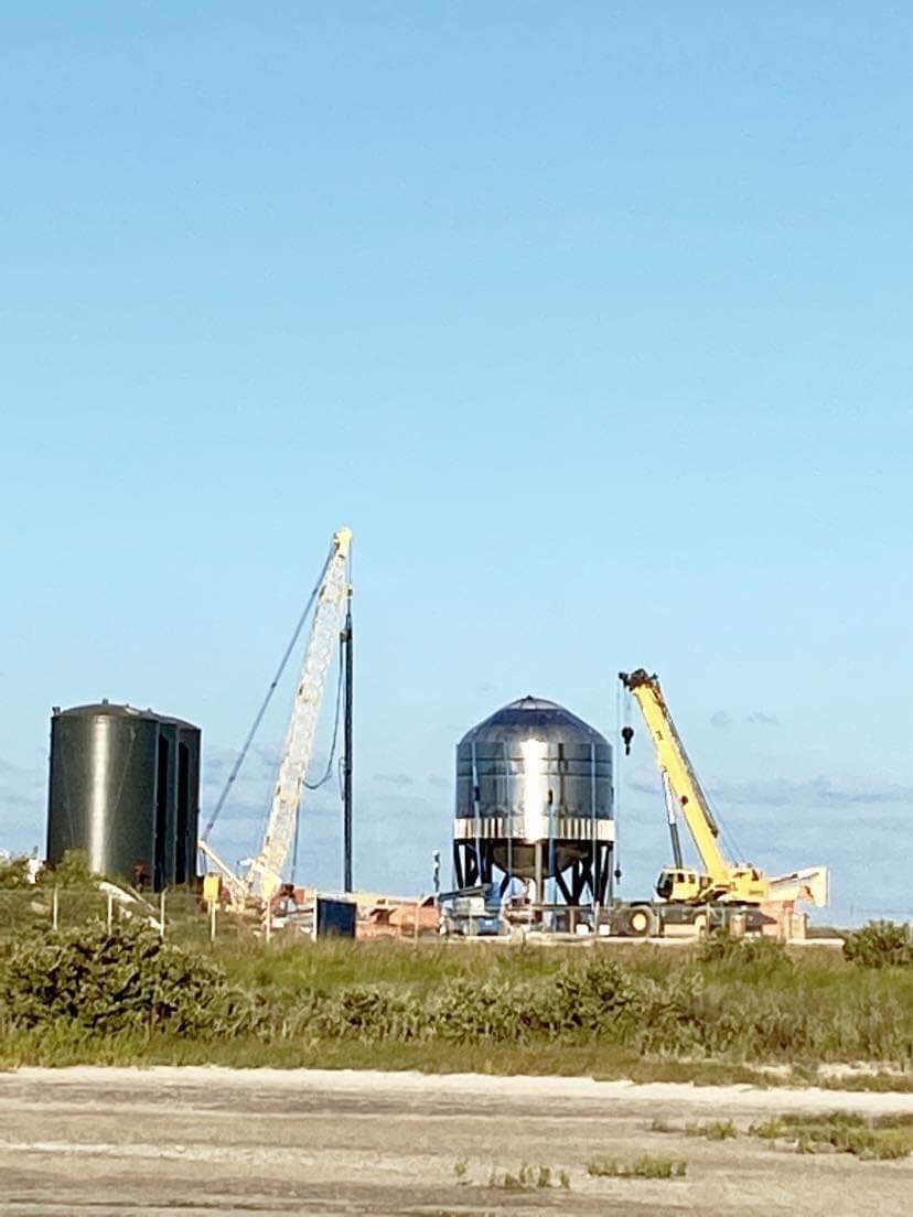 A stainless steel Starship test tank on a test stand between two cranes, with dark storage tanks to the left and brush in the foreground