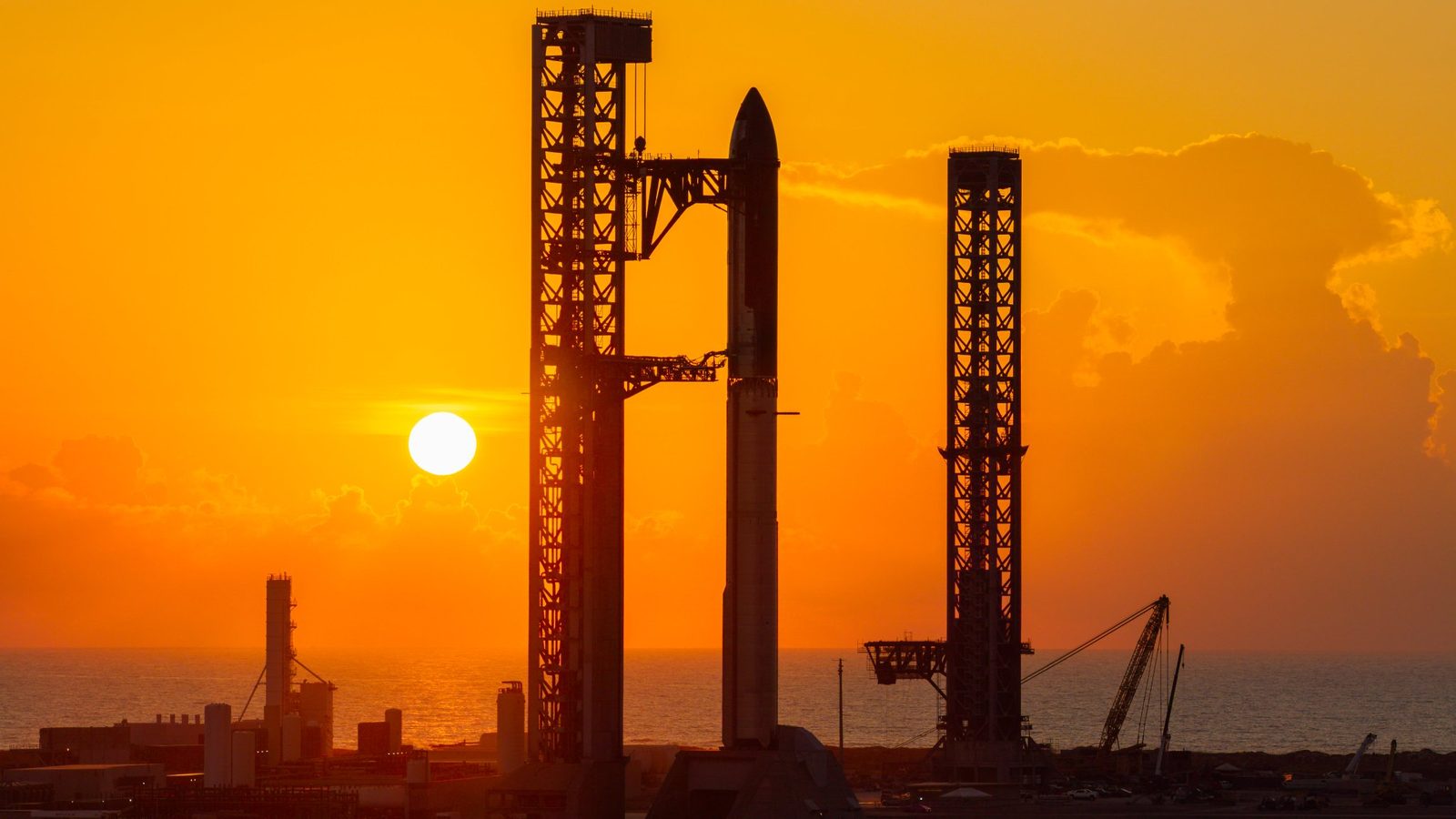 The Starbase launch tower and a Starship stack in silhouette against an orange sunrise over the Gulf