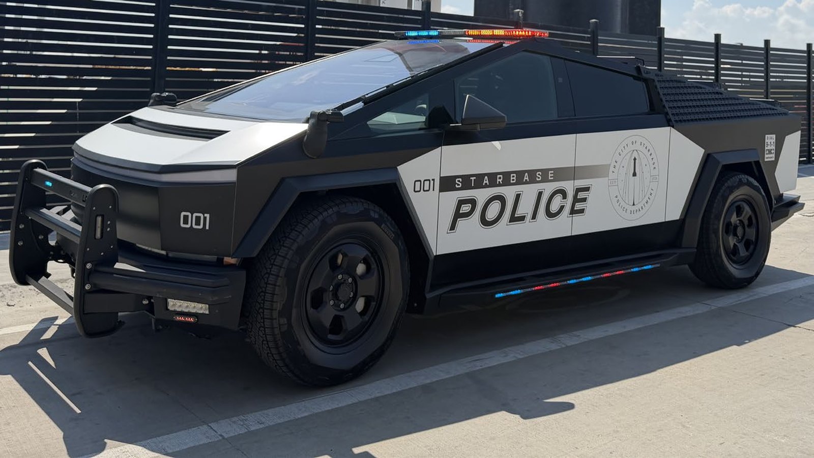 A Starbase police Cybertruck, unit 001, in a black and white police wrap with a push bumper and roof light bar, parked beside a slatted black fence