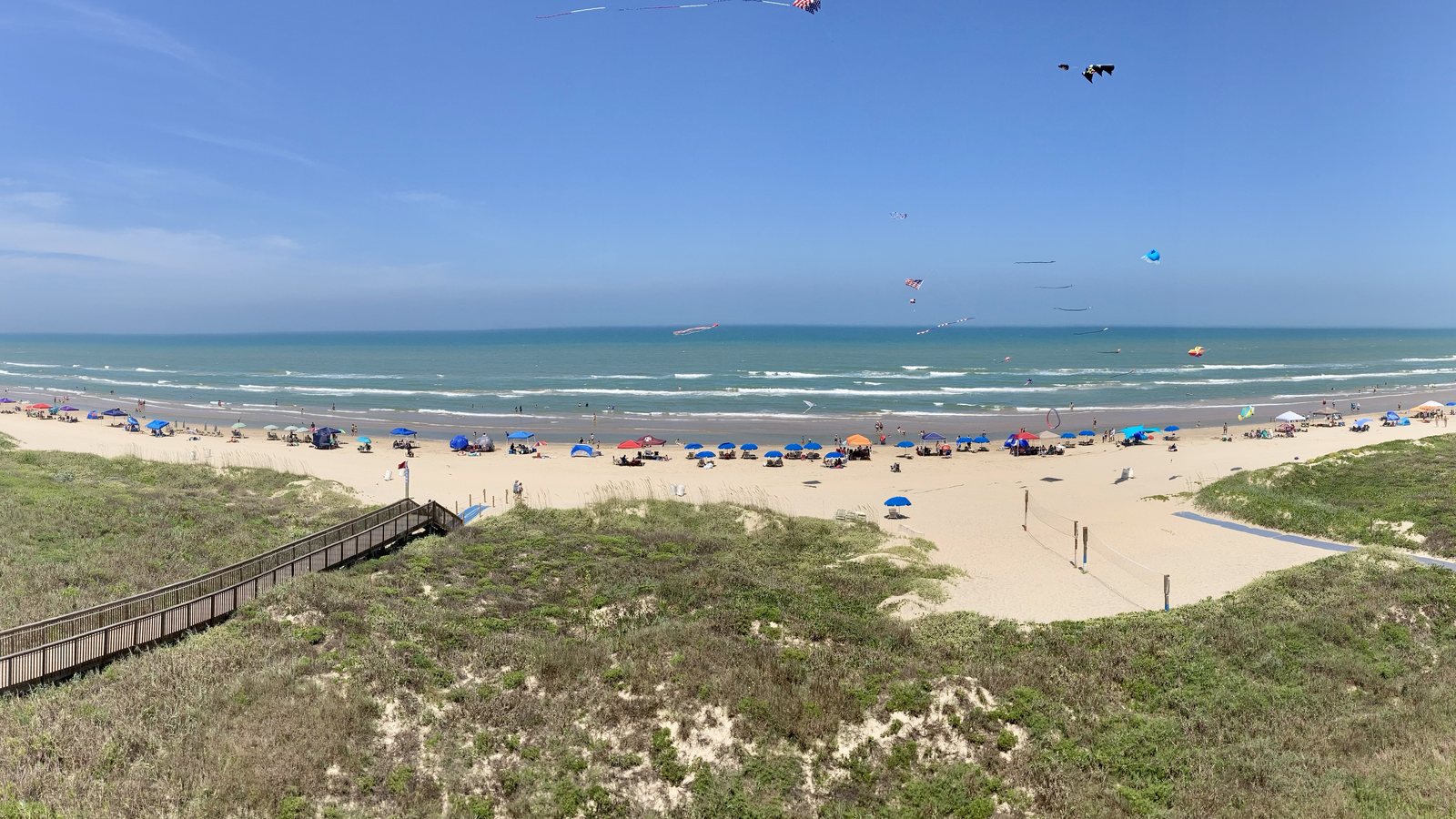 A wooden dune walkover leads down to a wide South Padre Island beach lined with umbrellas, with kites flying over the Gulf on a clear day