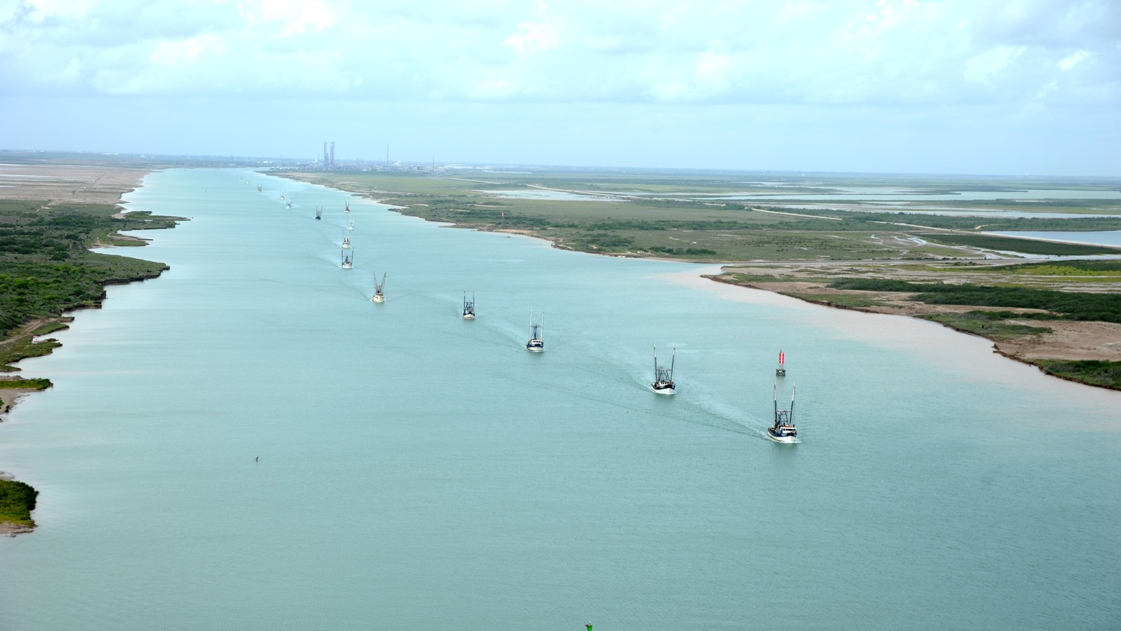 Shrimp boats heading single file down the wide Brownsville Ship Channel, with flat green marsh on both banks