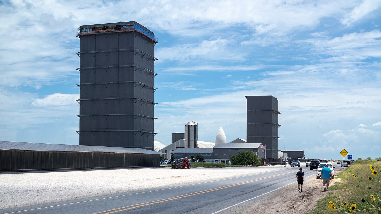 Highway 4 running past a tall gray SpaceX building and hangars at Starbase under a partly cloudy sky